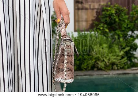 Fashionable woman holding luxury snakeskin python bag. Elegant outfit. Close up of purse in hands of stylish lady. Model posing near the swimming pool.