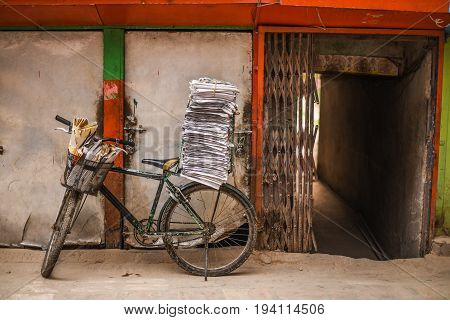 old bicycle with stack of newspapers near rustic wall and opened entrance gate of building urban street scene of Katmandu Nepal