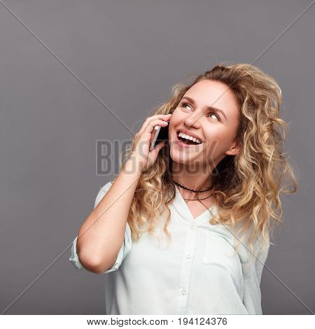 Blonde young curly haired woman talking on phone smiling wearing white shirt with sleeves rolled up.