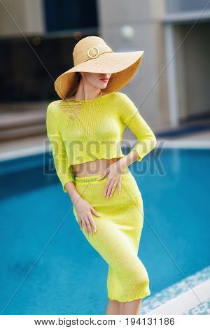 Beautiful woman in big hat posing next to the pool. Woman wears bright yellow outfit