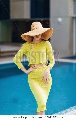 Beautiful woman in big hat posing next to the pool. Woman wears bright yellow outfit