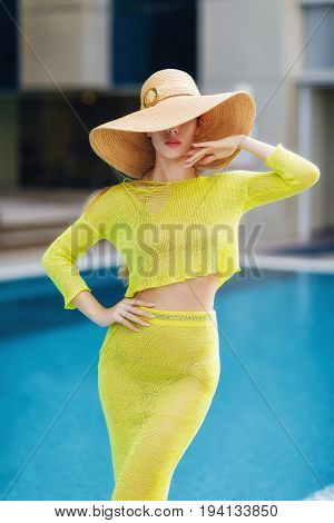 Beautiful woman in big hat posing next to the pool. Woman wears bright yellow outfit