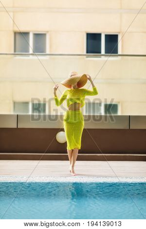Beautiful woman in big hat posing next to the pool. Woman wears bright yellow outfit