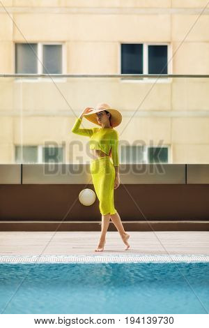 Beautiful woman in big hat posing next to the pool. Woman wears bright yellow outfit