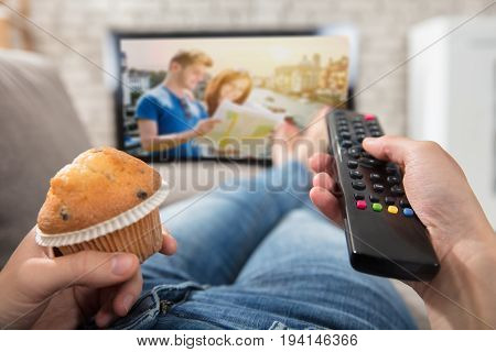Woman Holding Cupcake And Using Remote Control To Change The Channel While Watching Television