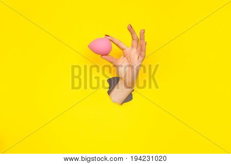 Crop female hand holding beauty blender in hole of yellow paper.