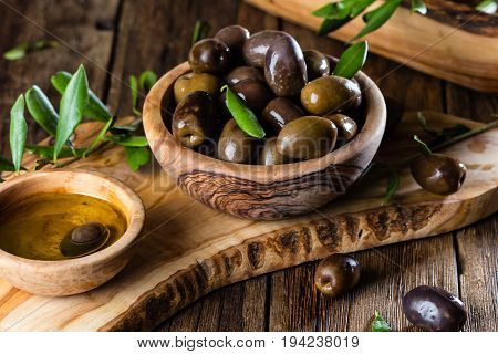 Olives and olive oil in olive wooden bowls on olive wooden cutting board, olive tree branch on wooden rustic background
