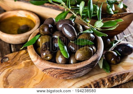 Olives and olive oil in olive wooden bowls on olive wooden cutting board, olive tree branch on wooden rustic background