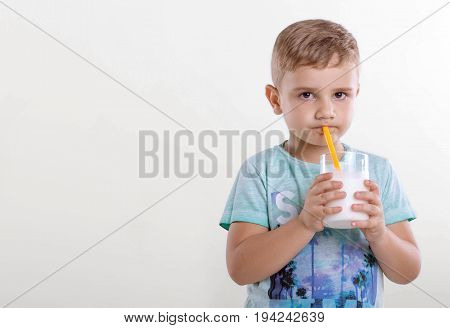 Portrait of a nice little boy in a blue T-shirt,  with a glass of milk and yellow straw. A cute little kid drinking a milk from a yellow straw on gray background.