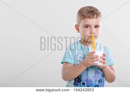 A cute little child with a glass full of fresh milk. Pretty little boy is drinking a glass full of milk from yellow straw on a gray blackground. Beautiful little boy is holding glass with milk shake.