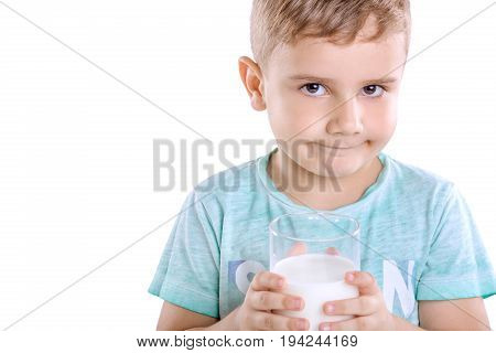 Curious little boy in a blue t-shirt is holding a full glass of milk isolated on a white background. Close-up portrait of happy little boy drinking fresh milk .