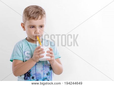 Pretty little boy in the blue t-shirt is drinking milk from a yellow straw on a gray background. Beautiful child with glass of milk on gray background. Healthy eating little boy is drinking milk from a yellow straw.