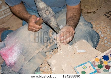 A moroccan craftsman is sitting on the floor making mosaics out of ceramic using a hammer.
