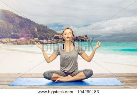 fitness, people and healthy lifestyle concept - woman doing yoga meditation in lotus pose on mat over exotic tropical beach background