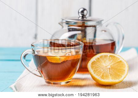 Glass cup of tea with lemon and glass teapot on blue wooden table.