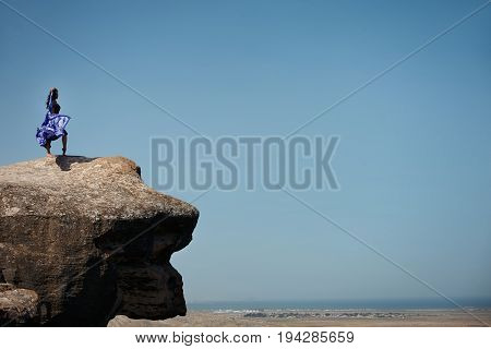 Young woman with colorful scarf enjoying nature on the mountains. The girl in black dress with long flying train stands on the top of the rock on beautiful landscape.