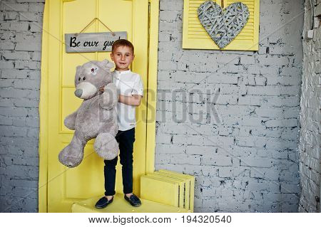 Portrait Of A Little Cute Boy Officially Dressed Posing With A Teddy Bear On Yellow Boxes In Grey Ro