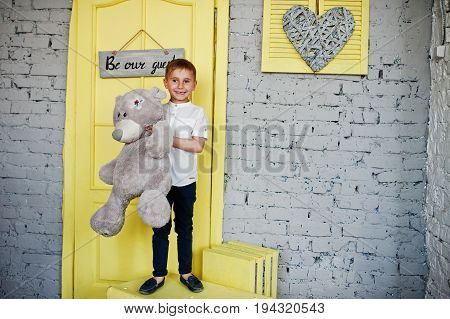 Portrait Of A Little Cute Boy Officially Dressed Posing With A Teddy Bear On Yellow Boxes In Grey Ro