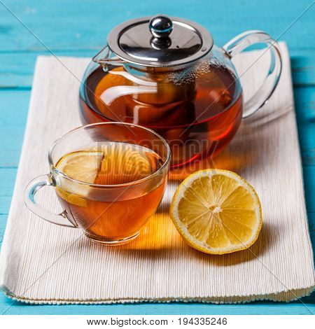 Glass cup of tea with lemon and glass teapot on blue wooden table.