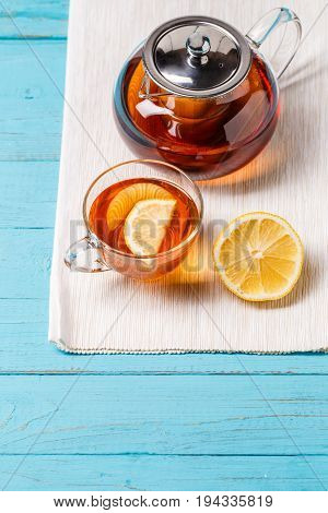 Glass cup of tea with lemon and glass teapot on blue wooden table.