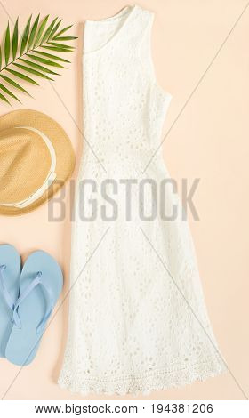 Summer fashion summer outfit on cream background. White lace dress blue flip flops and straw hat. Flat lay top view