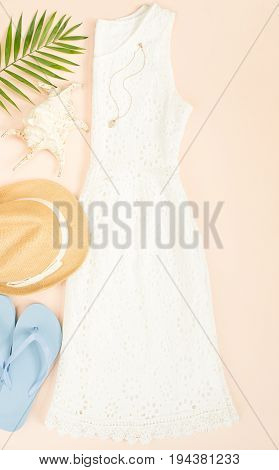 Summer fashion summer outfit on cream background. White lace dress blue flip flops and straw hat. Flat lay top view