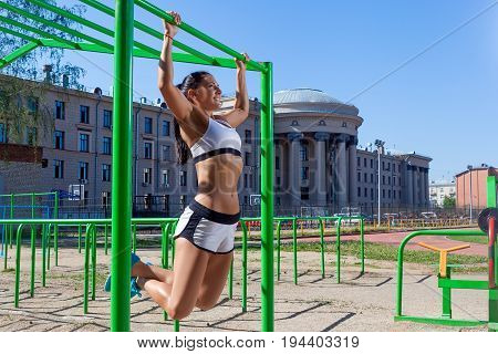 Young athletic brunette woman doing pull-up on a sports horizontal bar in a stadium on a summer day