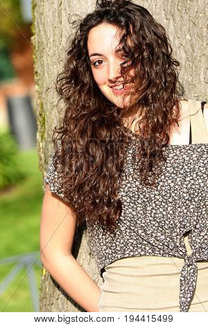 Curly, beautiful young girl with nice smile. Beautiful and young smiling woman, long hair style. A young italian teenager stands near a tree and smiles. Long wavy hair.