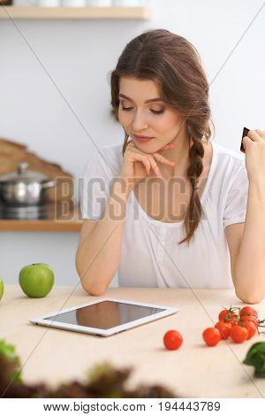 Young  woman looking for a new recipe for  cooking in a kitchen. Housewife is making online shopping by tablet computer and credit card.