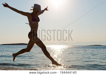 Young happy woman in swimsuit on the beach running and jumping on the seashore. Shot taken in backlit.