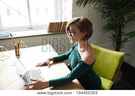 Portrait of serious and pretty woman in green drees working on testing new web site of fashion company. Concept of working online in the internet. Attractive manager sitting at the desk with laptop.