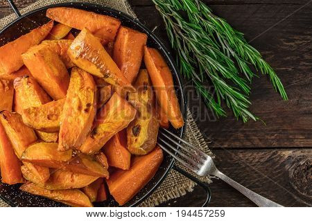 An overhead photo of roasted sweet potatoes in a pan, shot from above on dark rustic wooden textures with rosemary branches, with a place for text and a fork