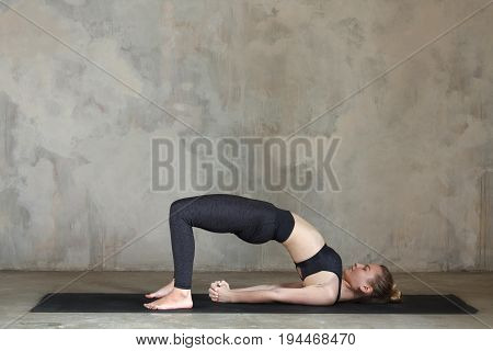 Young Woman Practicing Bridge, Setu Bandhasana Yoga Pose Against Texturized Wall / Urban Background