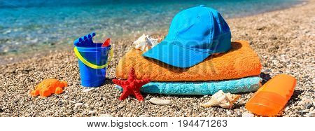 Beach towels, cap, children's toys, seashells and sunscreen on the beach on a background of a sea and mountains, summer family vacation by the sea concept. Horizontal. Close-up.