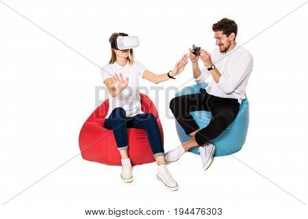 Smiling friends experiencing virtual reality glasses seated on beanbags isolated on white background. Experiencing virtual gaming adventure.