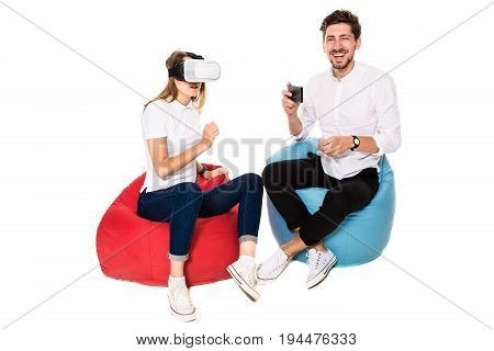 Smiling friends experiencing virtual reality glasses seated on beanbags isolated on white background. Experiencing virtual gaming adventure.