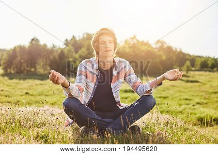 Outdoor Portrait Of Teenger Sitting On Green Grass Crossed Hands Closing His Eyes Meditating Trying