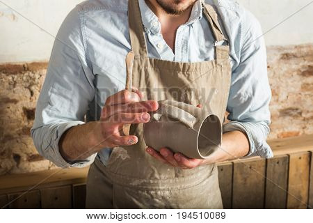 pottery, stoneware, ceramics art concept - closeup moisturizing of an unfinished fireclay product with water to bond the parts, craftsman stands at a workshop with a clay cup and connected handle