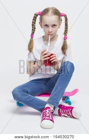 Kids Concepts. Portrait of Little Caucasian Blond Girl with Long Pigtails Posing With Pink Pennyboard and Drinking Red Juice with Straw. Against White. Vertical Image