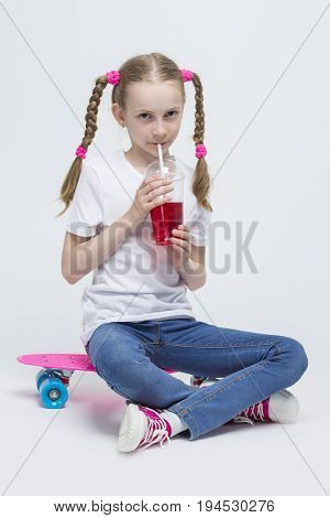 Kids Concepts. Portrait of Little Caucasian Blond Girl with Long Pigtails Posing With Pink Pennyboard and Drinking Red Juice with Straw. Against White. Vertical Shot