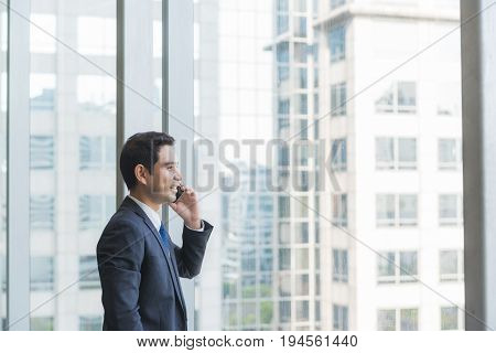 Mature and confident business executive looking looking out of large windows at a view of the city below from the top floor of an office building while talking on his mobile phone