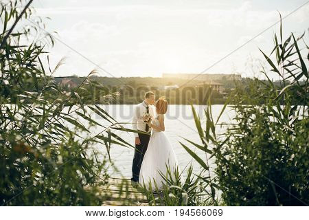 Elegant gentle stylish groom and bride near river or lake. Wedding couple in love. Woman in ehite dress man in white shirt and tie. With sunshine summer background.