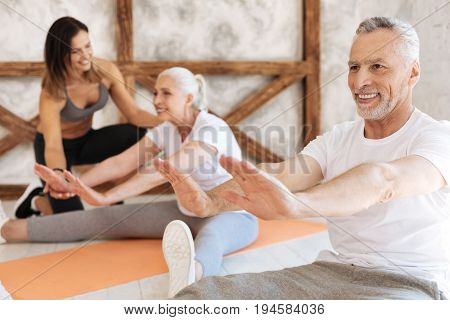 Do not hurry up. Positive male person keeping smile on his face while sitting on the floor on the foreground and doing exercises