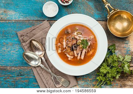 Soup with meat, olives, herbs, lemon in white bowl, black bread and spices on rustic wooden background, top view homemade food. Traditional Russian soup - solyanka