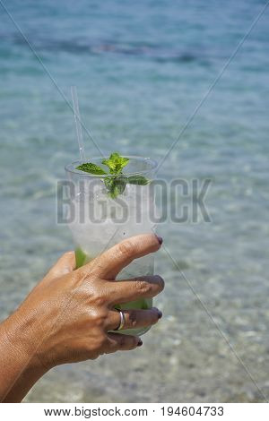 Cocktail drink in hand with blurred beach background.
Female hand holding an iced glass of mojito alcoholic cocktail with mint and sliced lime, on a sunny day.