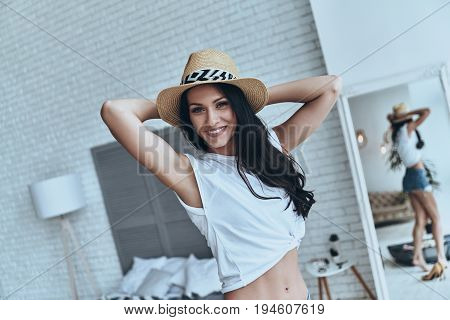 Ready foe vacation. Attractive young woman keeping hands behind head and smiling while standing in the bedroom at home