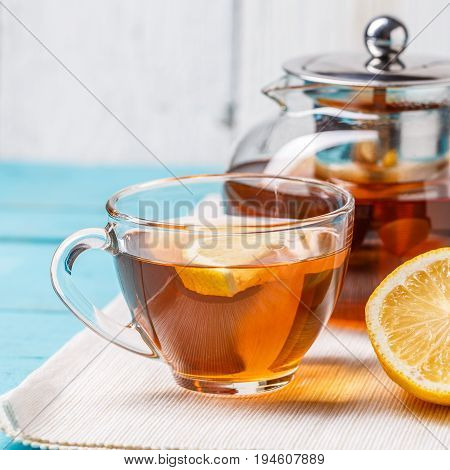 Glass cup of tea with lemon and glass teapot on blue wooden table.