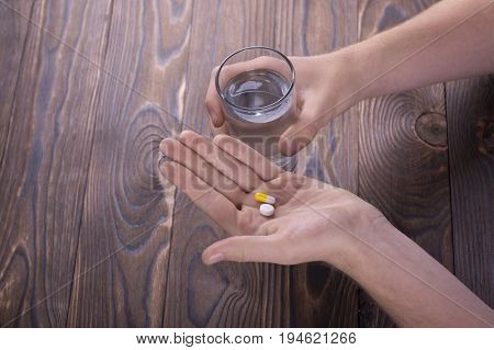 A man holds two pills and a glass of water in the hands of a wooden brown table. The guy drinks pills. Healthy generation. Healthy youth.