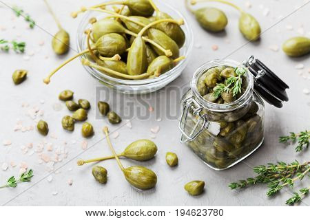 Mixed capers in jar and bowl on gray kitchen table from above.