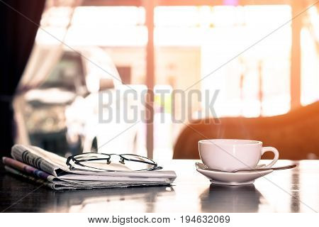 Closeup cup of hot cappuccino coffee with newspaper and spectacles on the wood table with blur coffee shop background-warm tone.